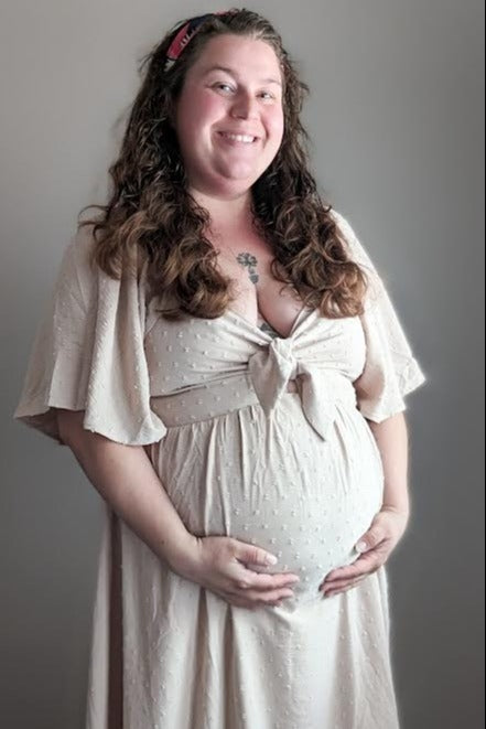 woman wearing a light beige maternity dress with a tie front, holding her baby bump and smiling