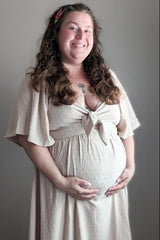 woman wearing a light beige maternity dress with a tie front, holding her baby bump and smiling