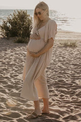 woman wearing a flowy beige maternity dress and holding her baby bump on a beach at sunset