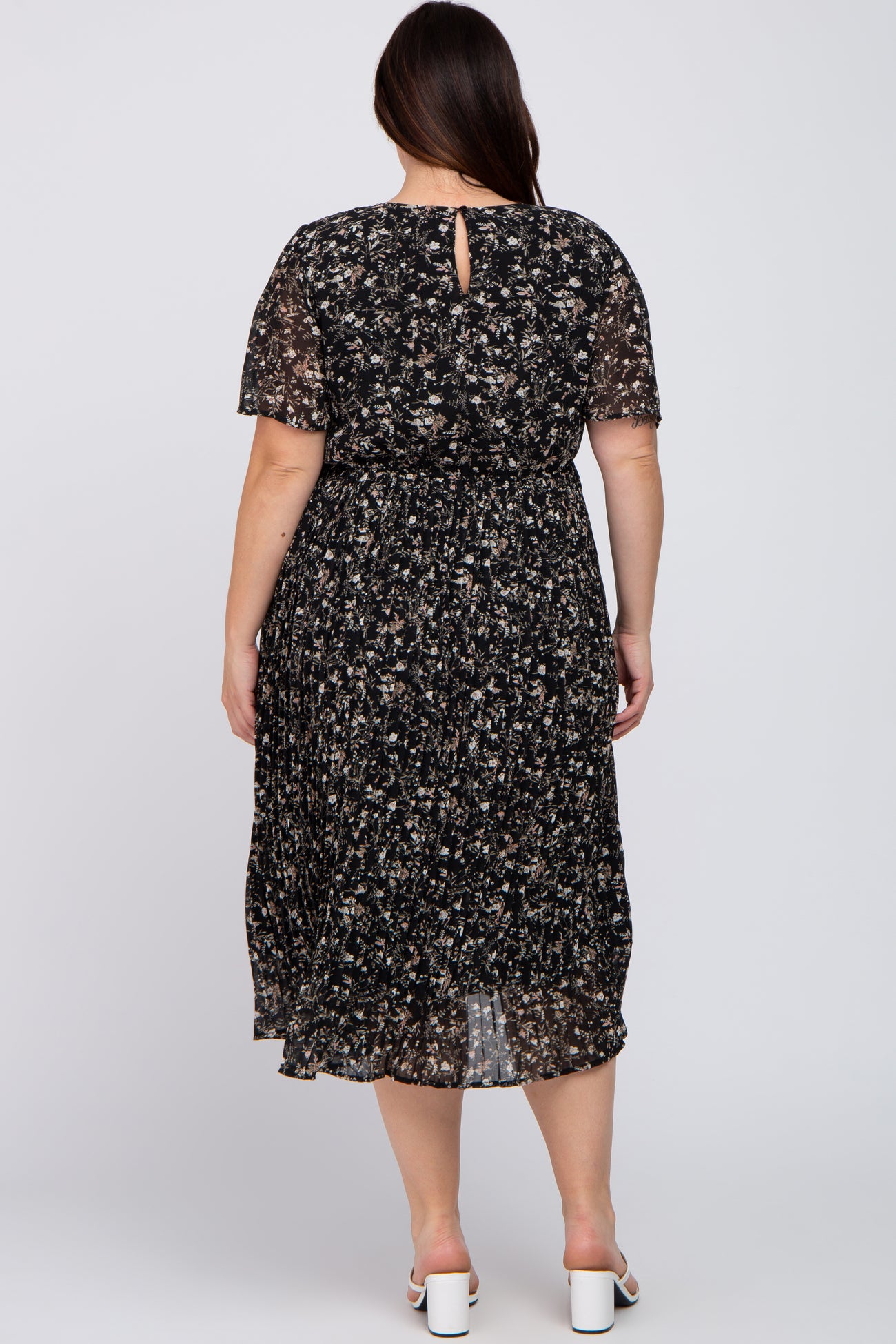 back view of woman wearing plus size black floral pleated midi dress with short sleeves and white heels