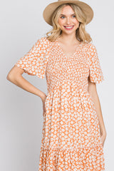 woman wearing an orange floral print short sleeve dress with a fitted bodice and tiered skirt, smiling and wearing a straw hat