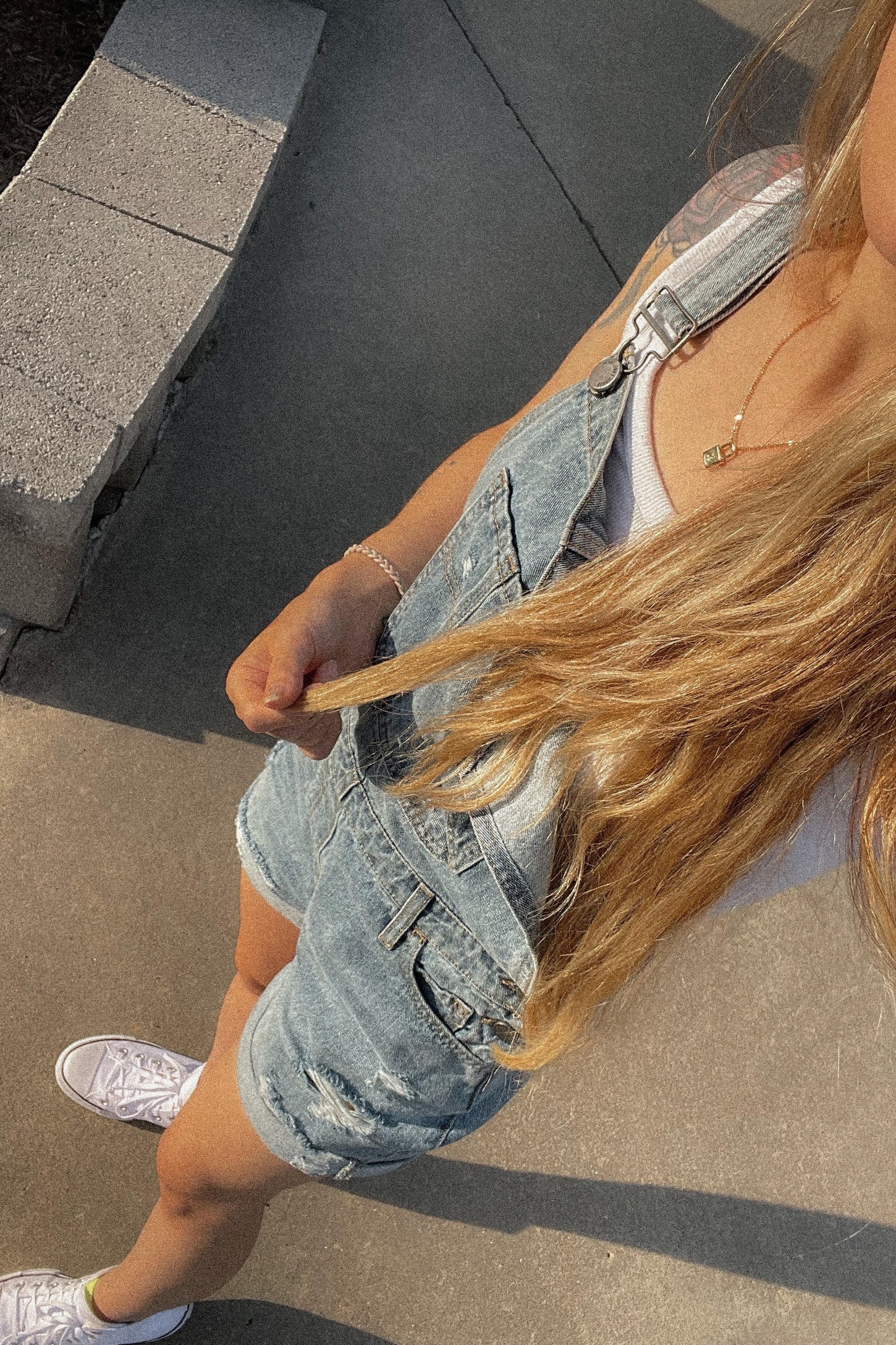 Woman wearing light wash denim overalls and white sneakers standing on a sidewalk in the sun