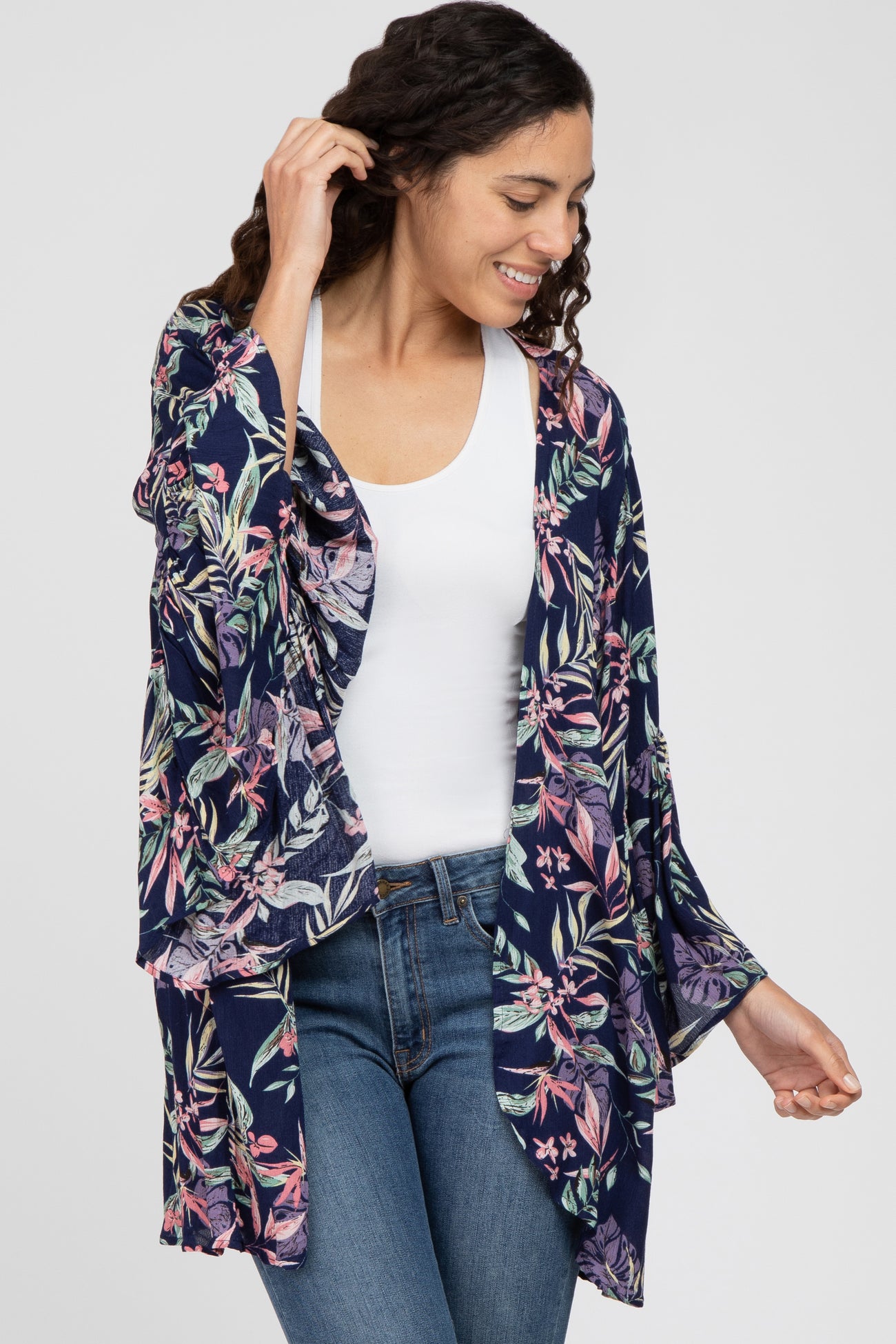 woman wearing a navy floral kimono cardigan over a white top and blue jeans, smiling and posing with one hand raised
