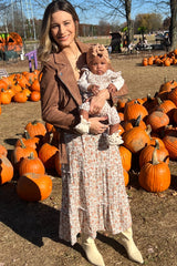 woman wearing a floral maxi dress and a brown jacket, holding a baby in a floral outfit among pumpkins