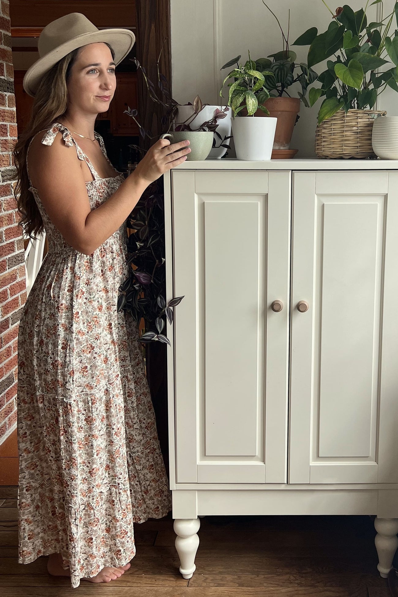 woman wearing a floral maxi dress with tie straps, standing by a cabinet and holding a plant pot