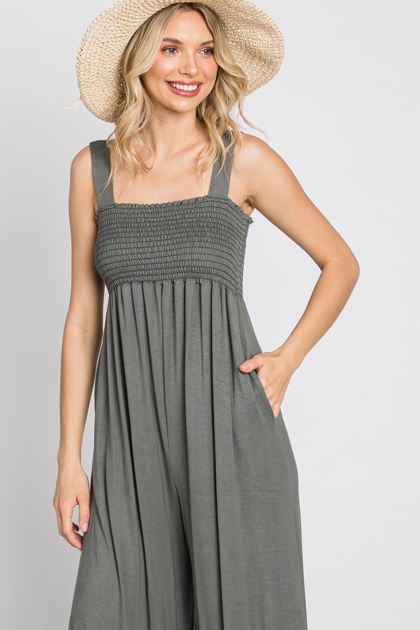 woman wearing a gray smocked jumpsuit with wide legs and pockets, holding a straw hat