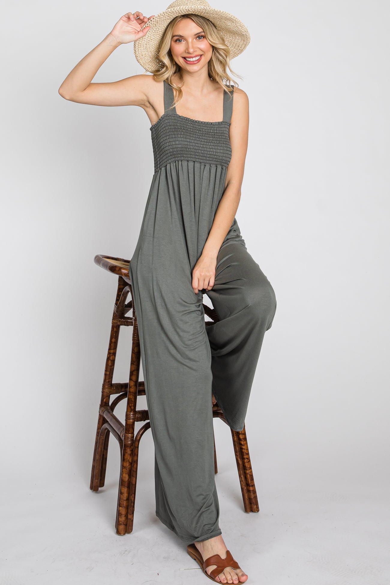 woman wearing a dark gray jumpsuit with a smocked bodice, sitting on a stool and wearing a wide-brimmed hat