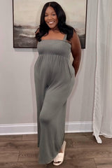 smiling woman wearing a sage green smocked jumpsuit and white sandals standing in a room