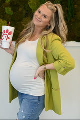 woman wearing a light green oversized blazer and a white maternity top, posing with a drink and showing her baby bump