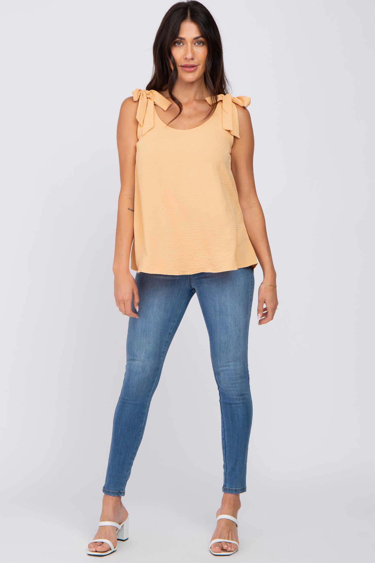 woman wearing a light yellow sleeveless top with bow tie straps and blue skinny jeans