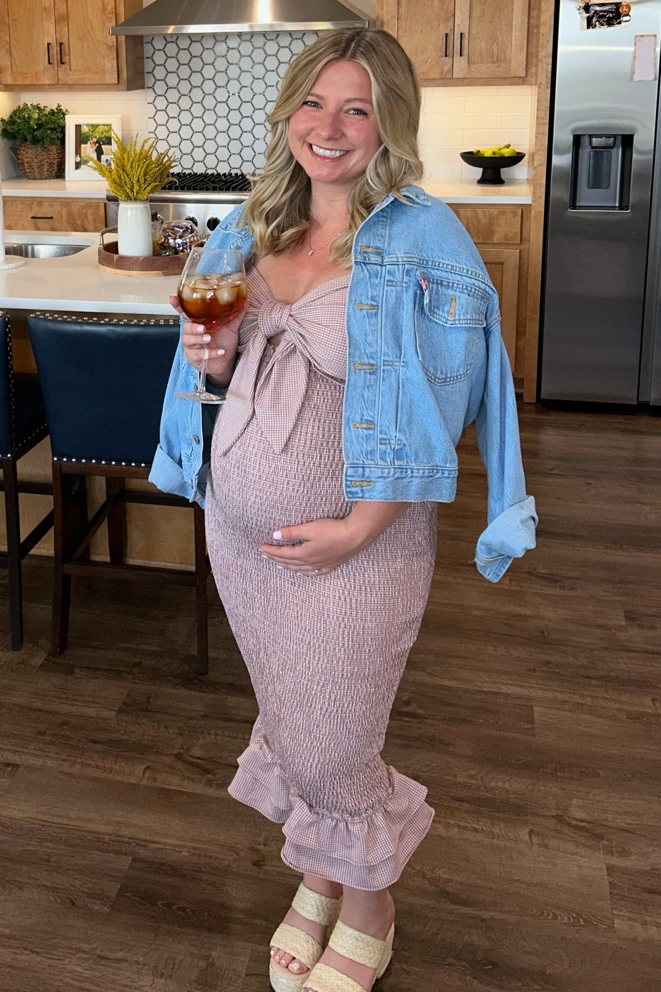 woman wearing a pink smocked maternity dress with a bow, blue denim jacket, and holding a drink, showcasing her baby bump