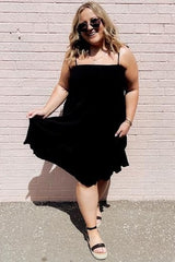 woman wearing a black spaghetti strap sundress and sunglasses standing against a pink brick wall