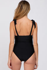 Back view of a woman wearing a black one-piece swimsuit with tie-shoulder straps against a white background