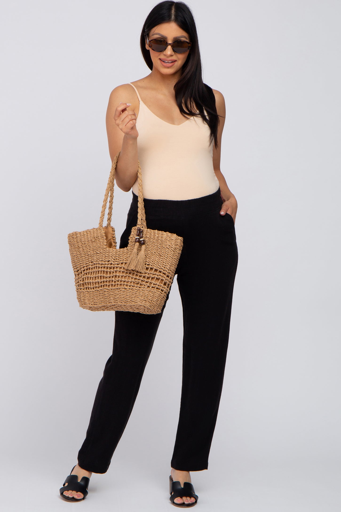 woman wearing a light nude maternity bodysuit with black wide-leg pants, holding a woven purse and wearing sunglasses