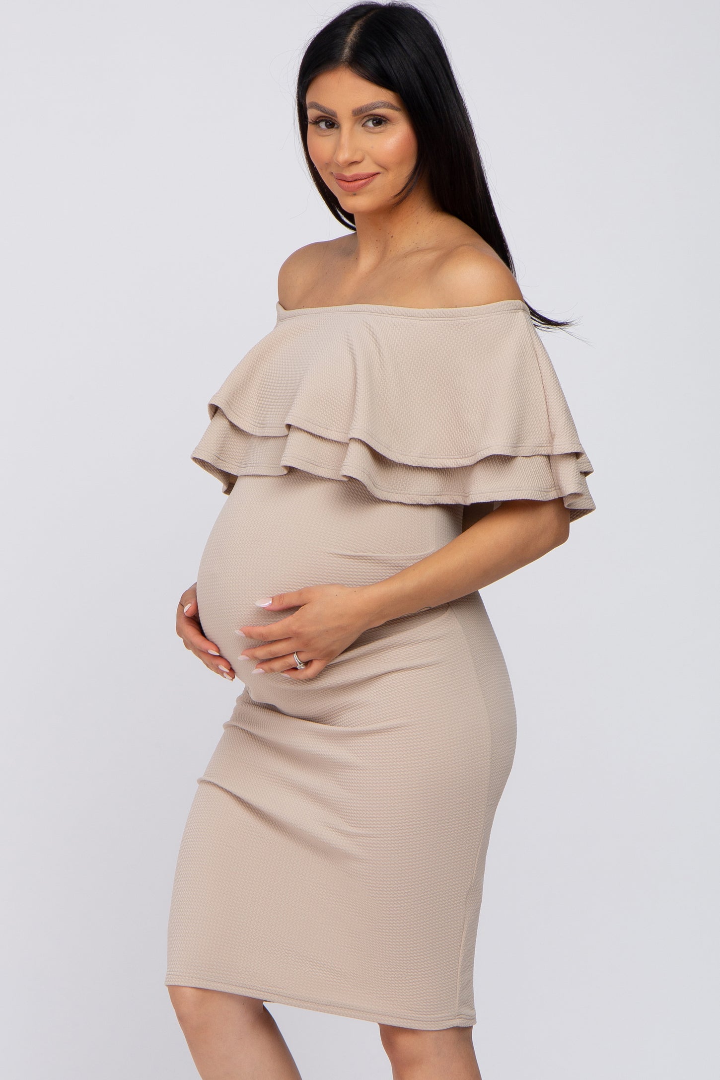 woman wearing a beige off-the-shoulder maternity dress with tiered ruffles and holding her baby bump