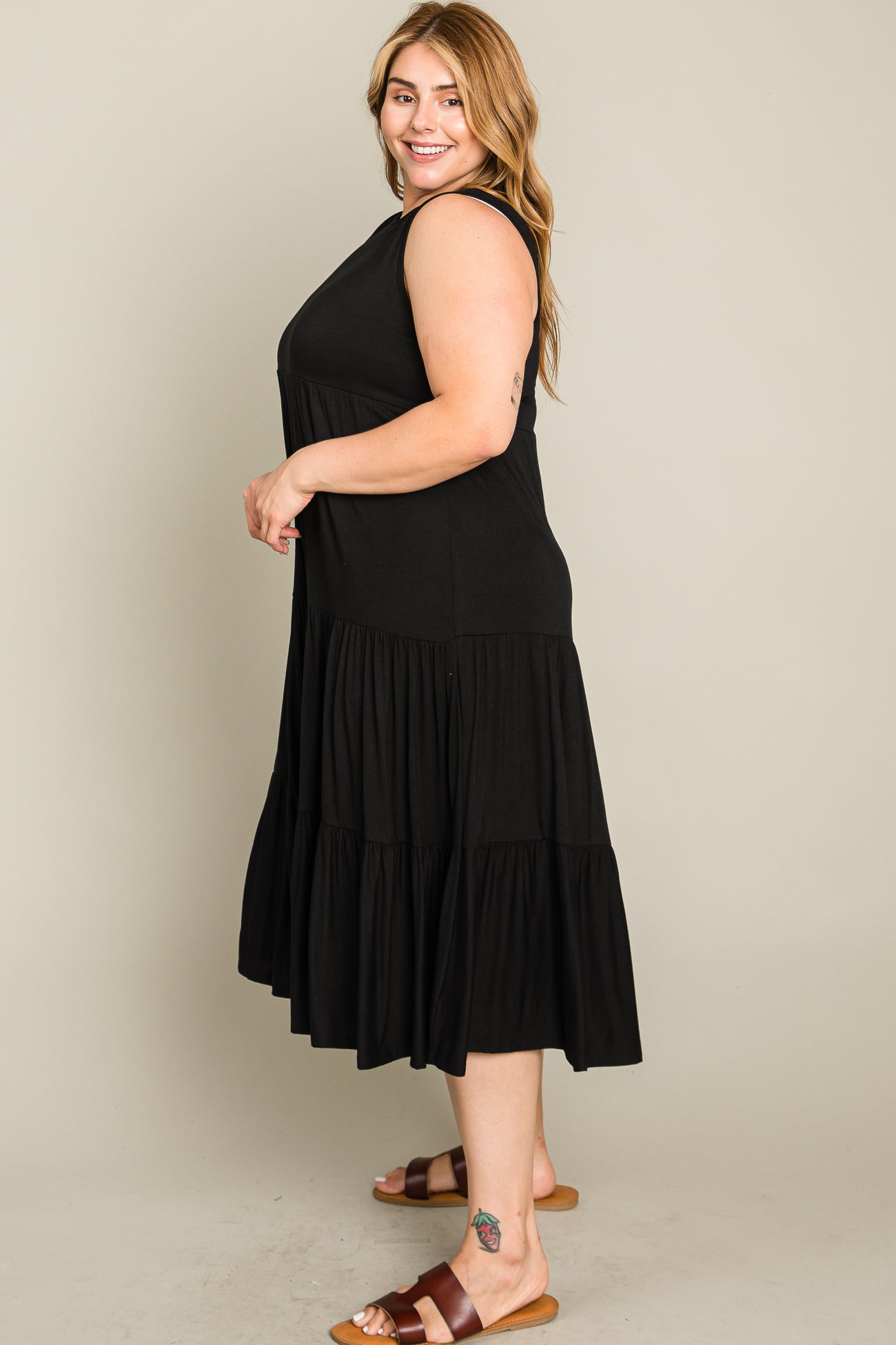 Smiling woman wearing a sleeveless black tiered midi dress and brown sandals against a neutral background