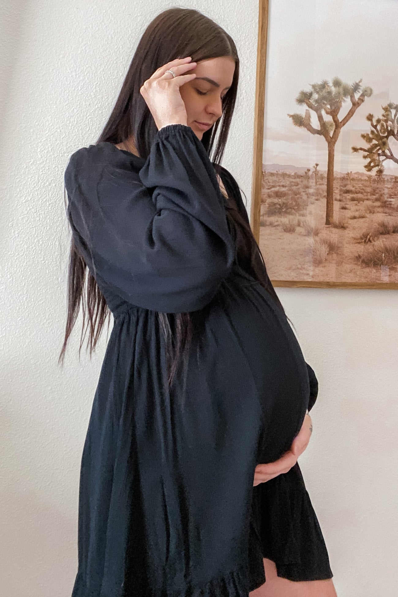 pregnant woman with long dark hair wearing a black long-sleeve maternity dress standing by a desert landscape painting