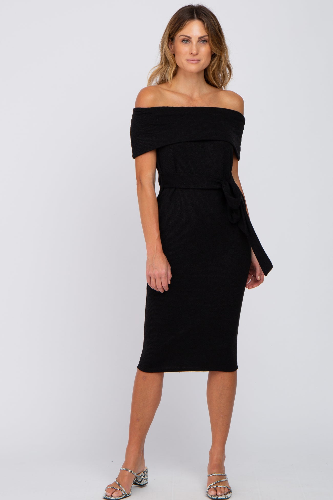 woman wearing a black off-the-shoulder fitted dress with a belted waist and a midi length, standing confidently