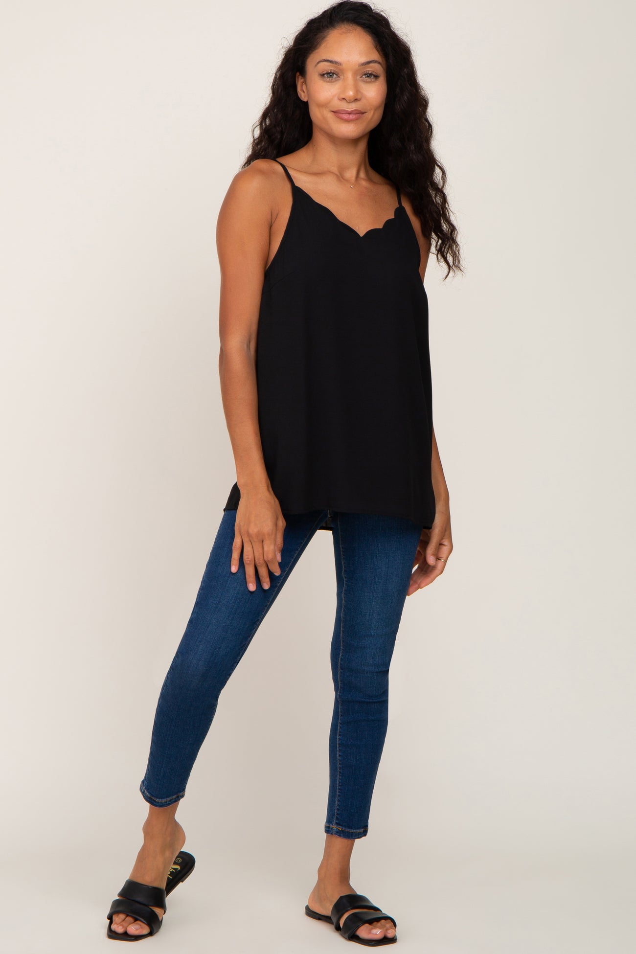 woman wearing a black scalloped camisole top and blue skinny jeans with black slide sandals