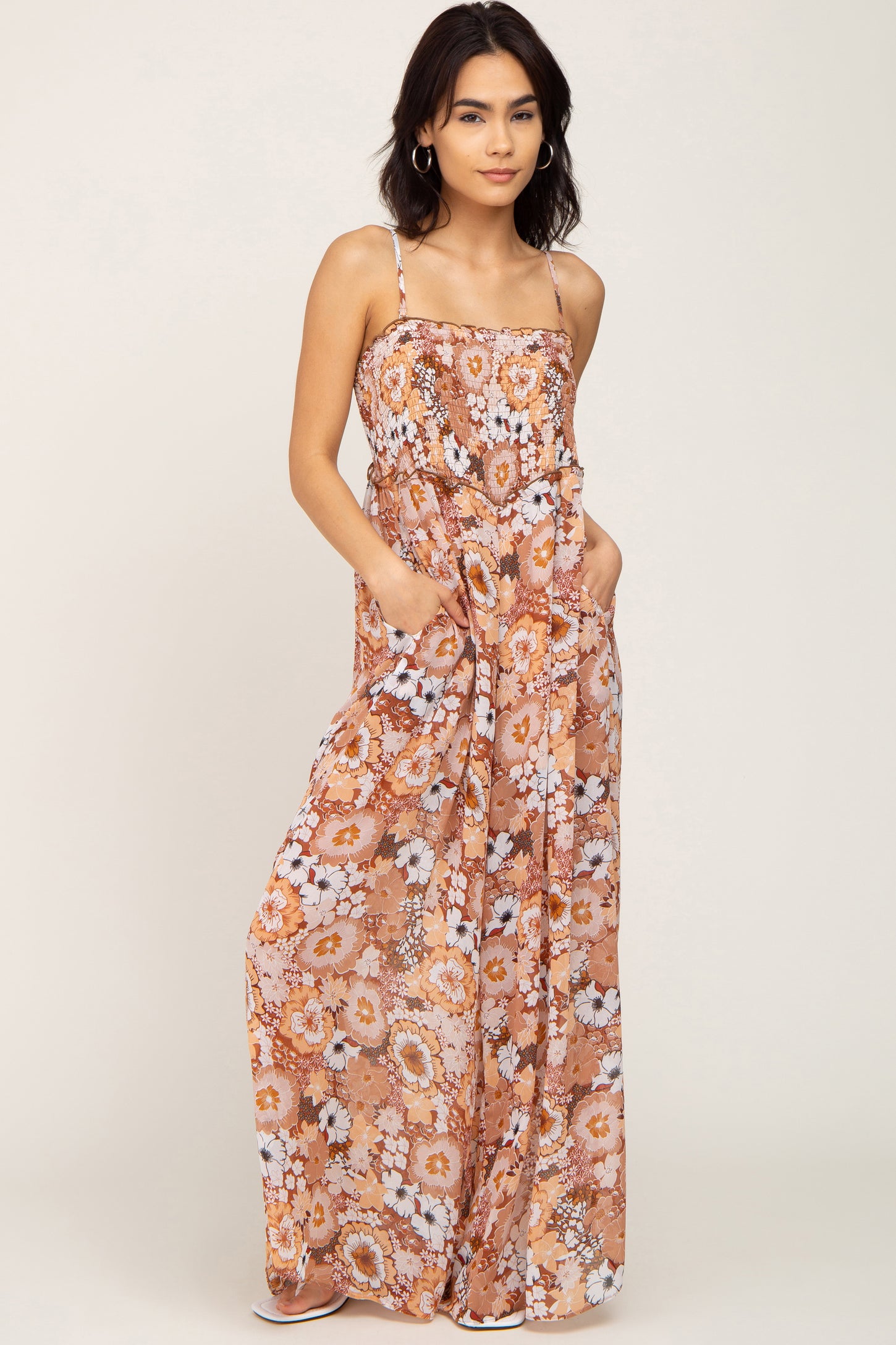 Woman wearing a floral print wide leg jumpsuit with spaghetti straps and pockets against a white background