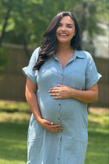 woman wearing a light blue maternity dress with buttons, holding her baby bump and smiling outdoors