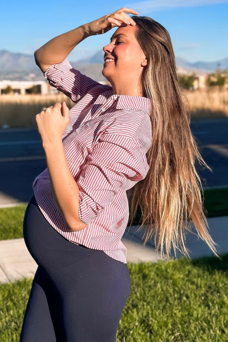 smiling pregnant woman in red striped shirt and navy leggings standing outdoors in sunlight