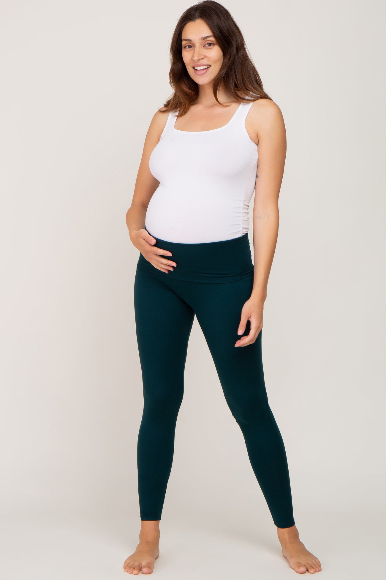 woman wearing a light pink maternity tank top and dark green maternity leggings, holding her baby bump