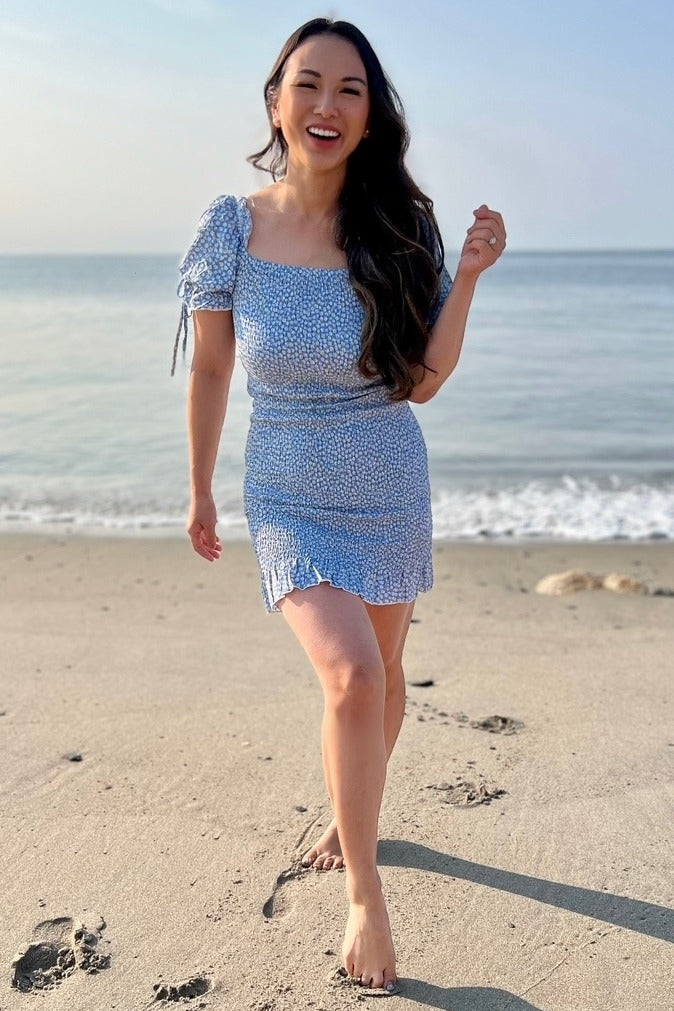 smiling woman in blue floral mini dress walking barefoot on a sandy beach with ocean waves in background