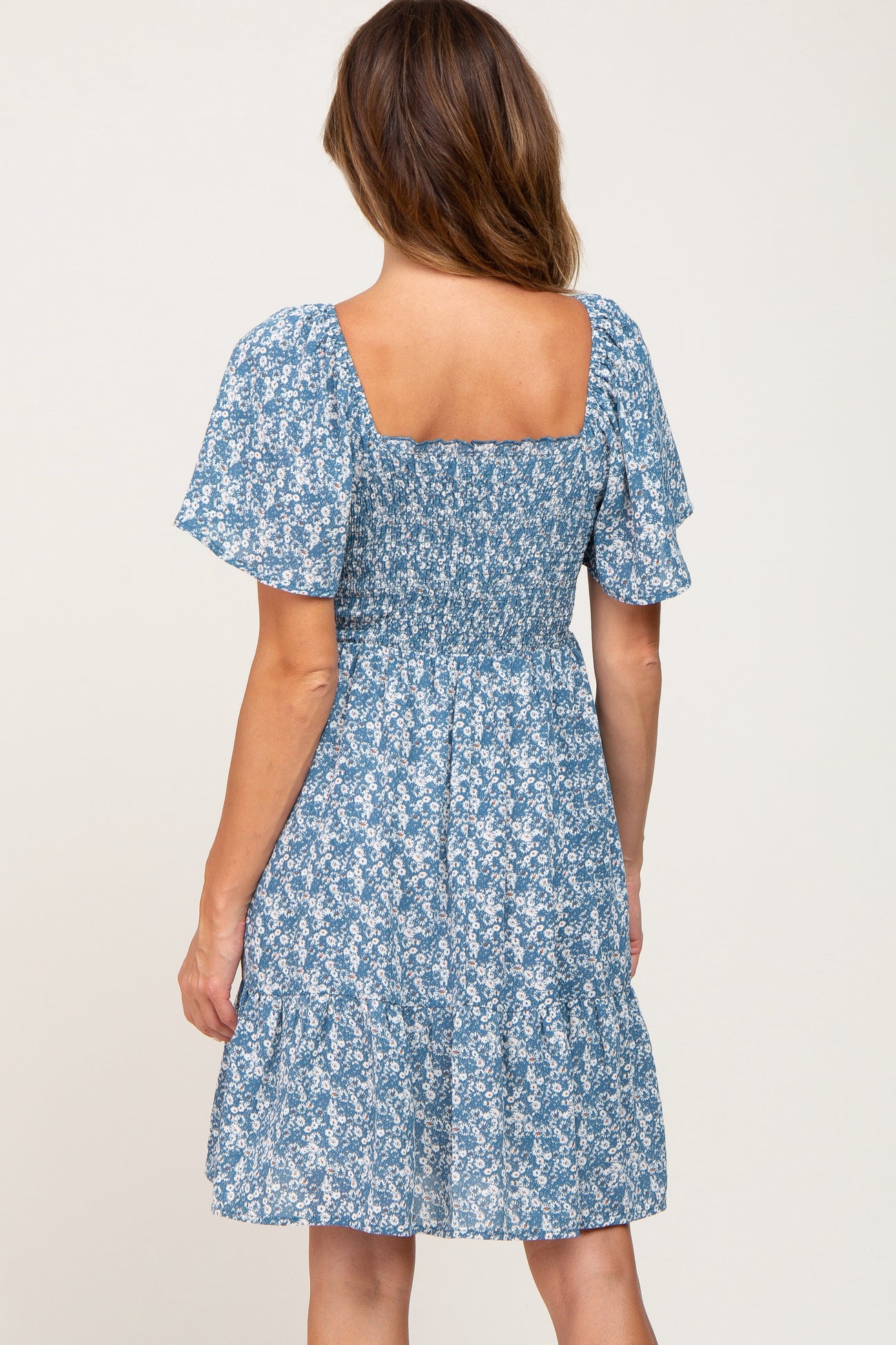 Back view of a woman wearing a blue floral smocked maternity dress with short flutter sleeves and a square neckline