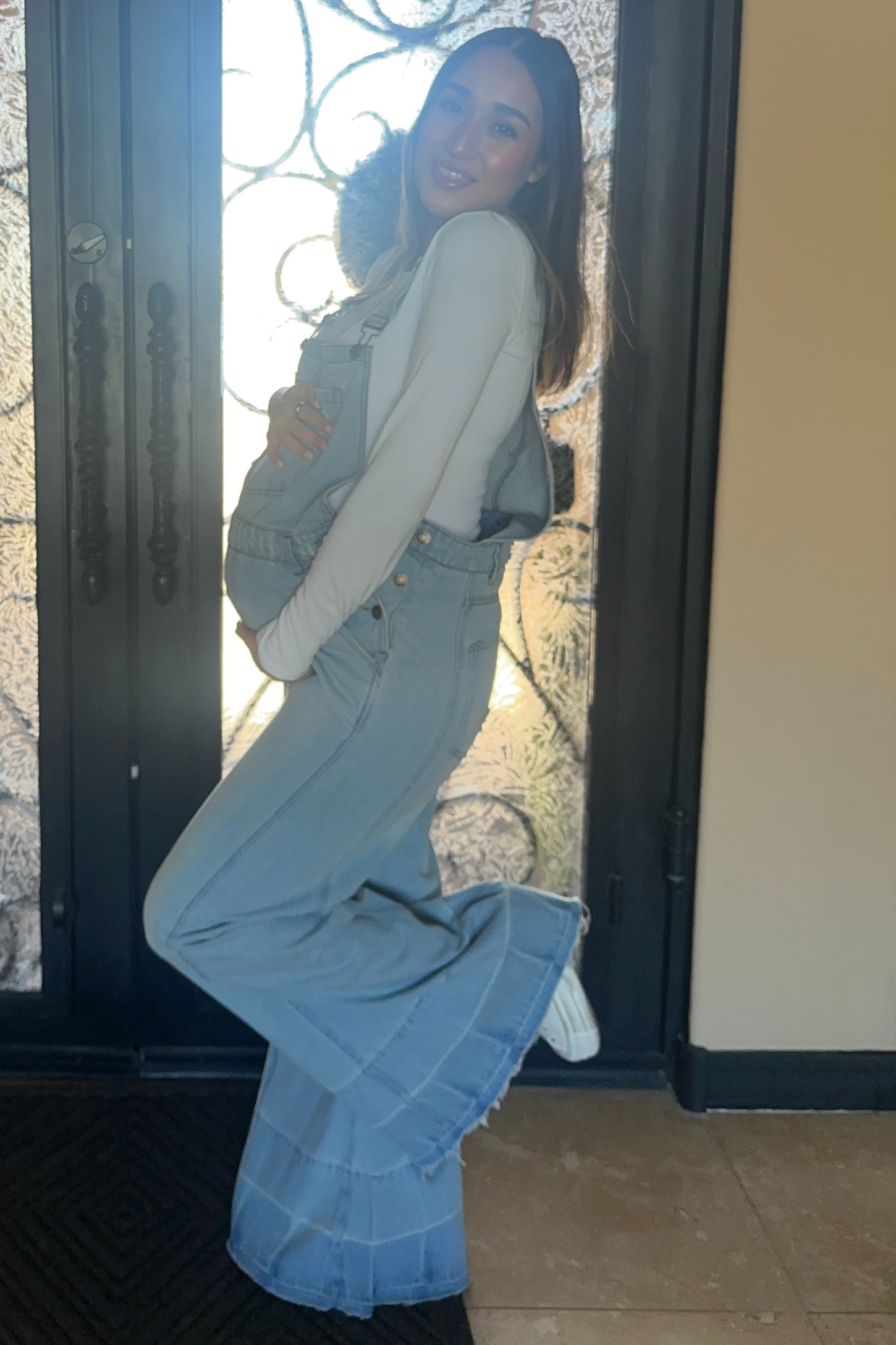 pregnant woman in light wash denim maternity overalls and white long sleeve shirt posing in front of a door