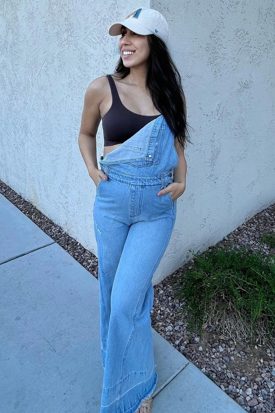 woman wearing light wash denim overalls and a brown crop top with a white baseball cap