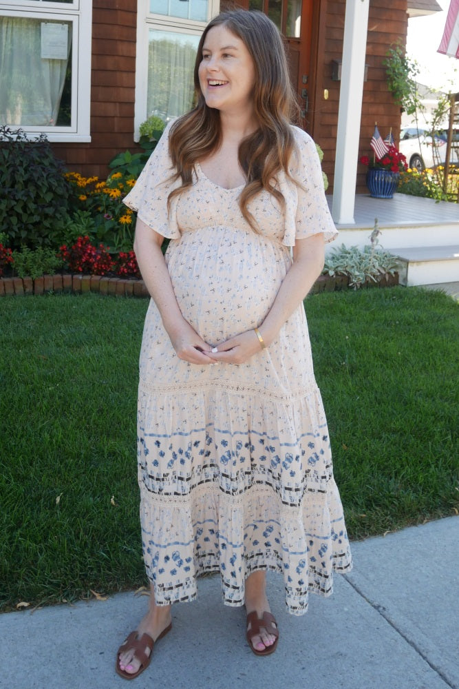 smiling pregnant woman wearing a floral maternity maxi dress standing on a sidewalk in front of a house