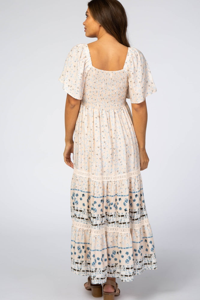 Back view of a woman wearing a cream floral smocked maternity maxi dress with flutter sleeves and tiered skirt