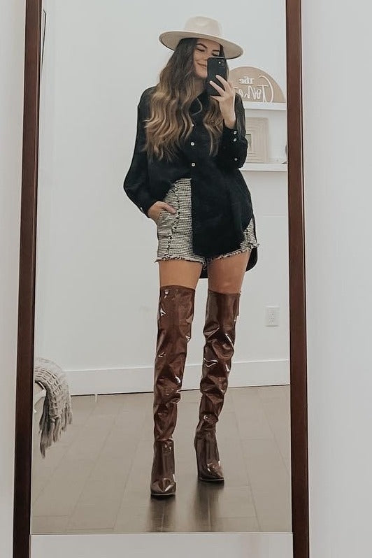 Woman in a black button-down shirt, tweed shorts, and brown patent leather over-the-knee boots taking a mirror selfie