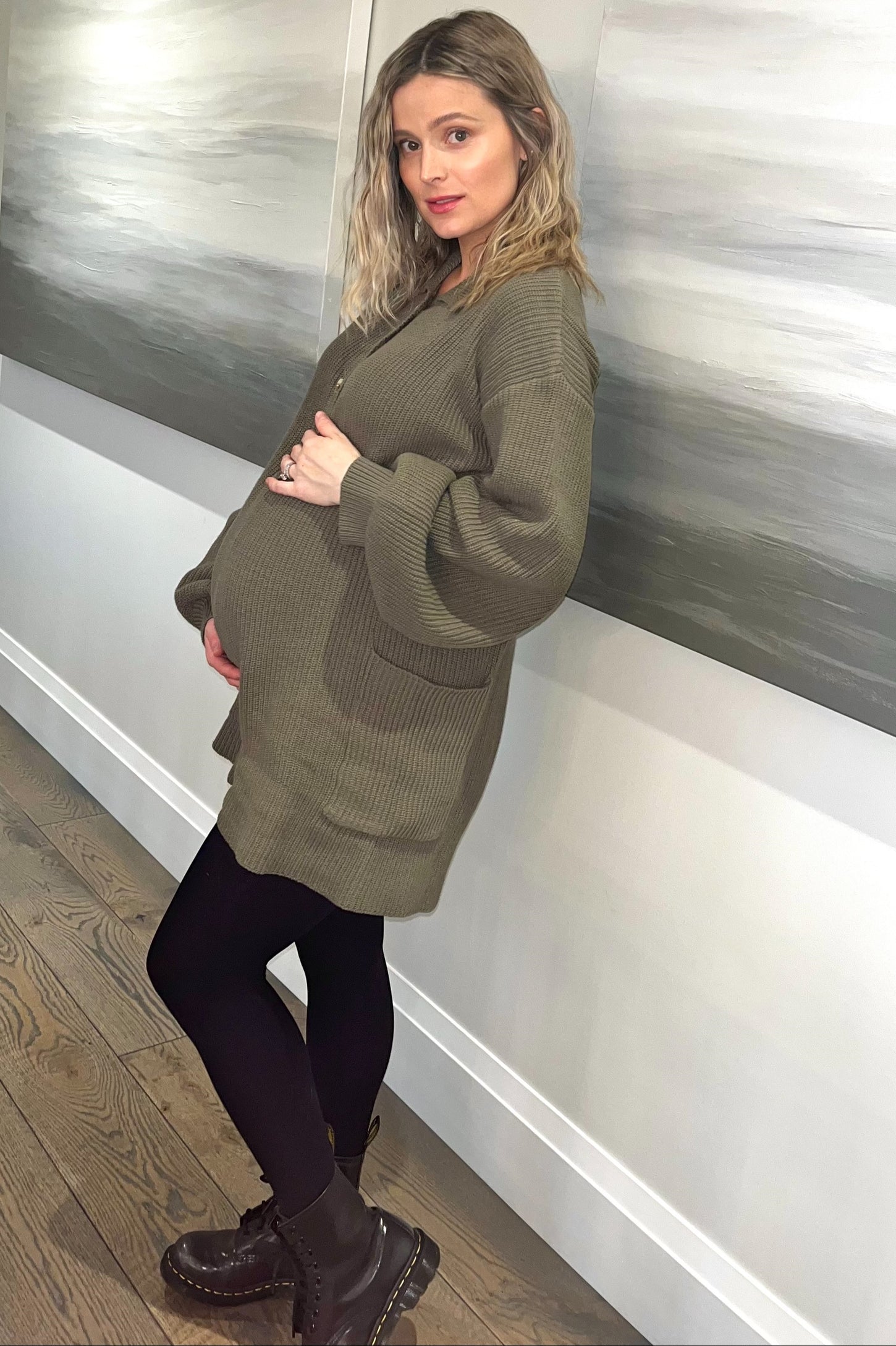 pregnant woman in olive green oversized cardigan and black leggings posing against a wall