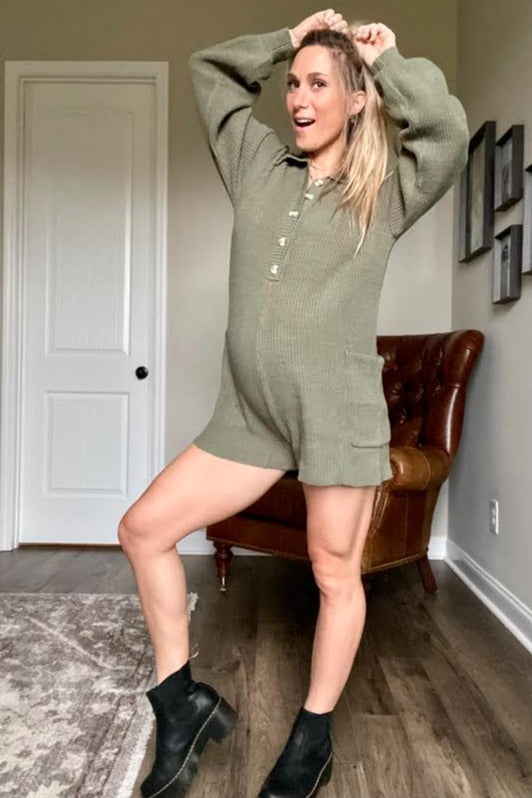 Pregnant woman wearing an olive green knit romper and black boots posing in a living room