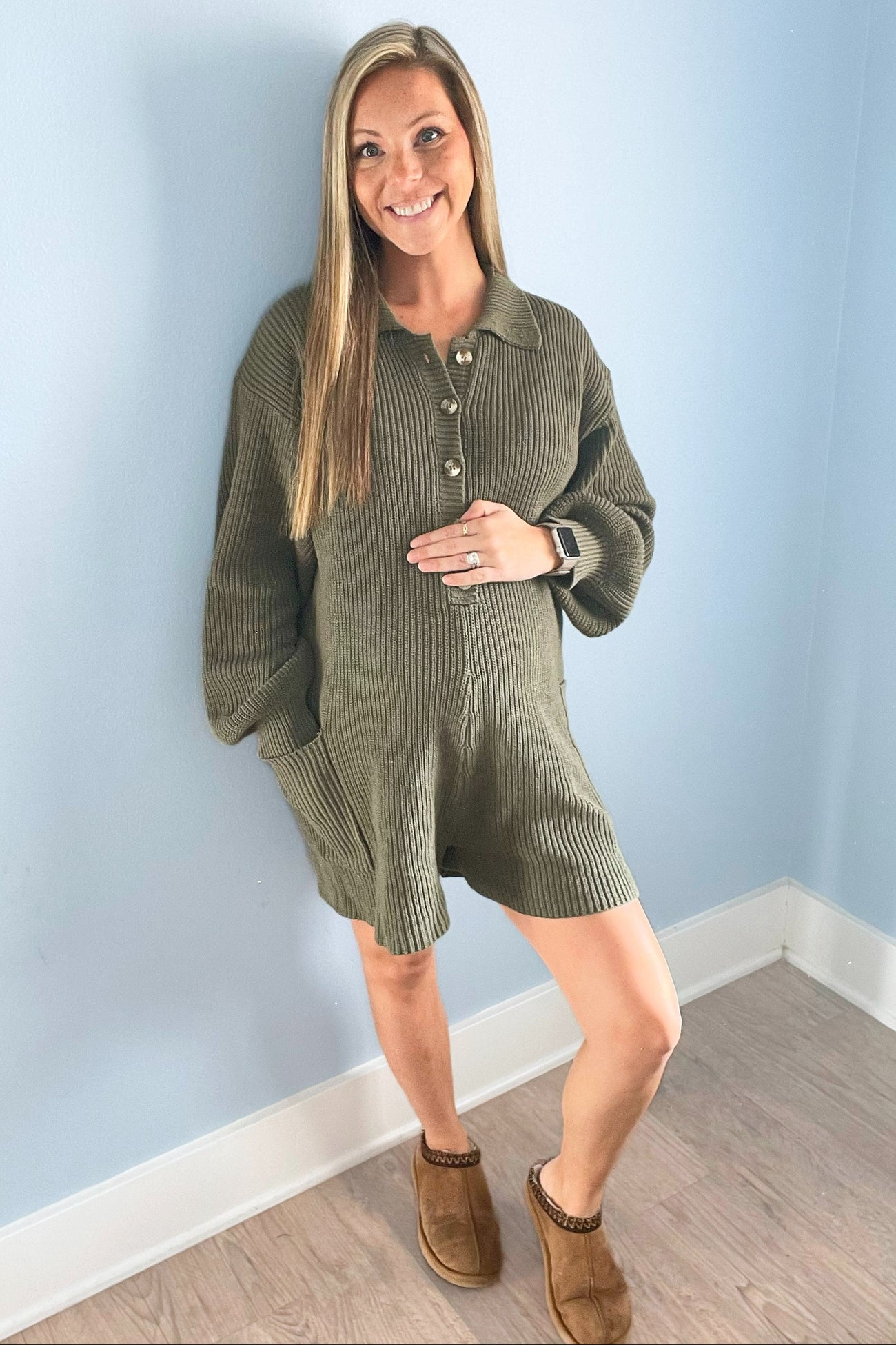 Smiling pregnant woman wearing an olive green ribbed knit maternity romper and brown slippers