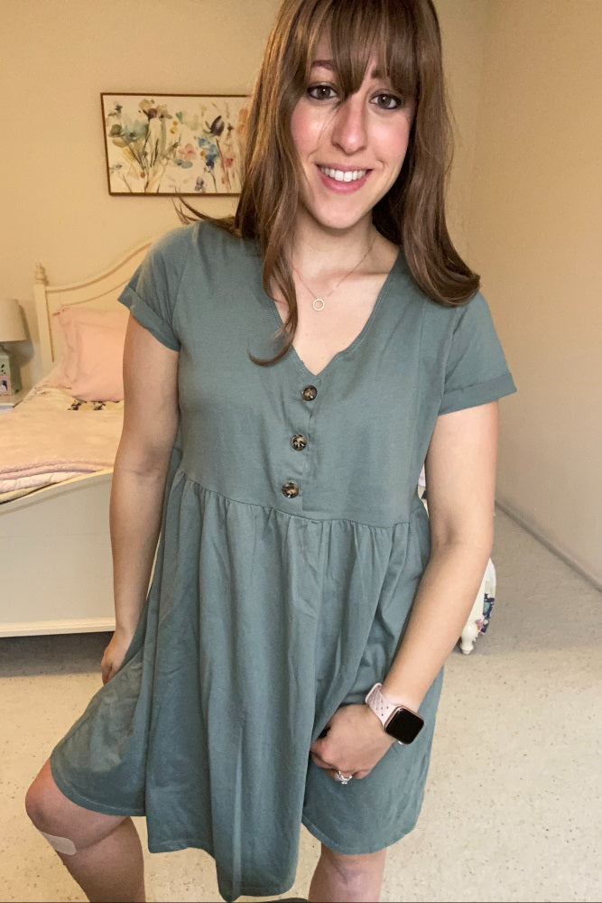 woman wearing a sage green short sleeve button front babydoll dress in a bedroom