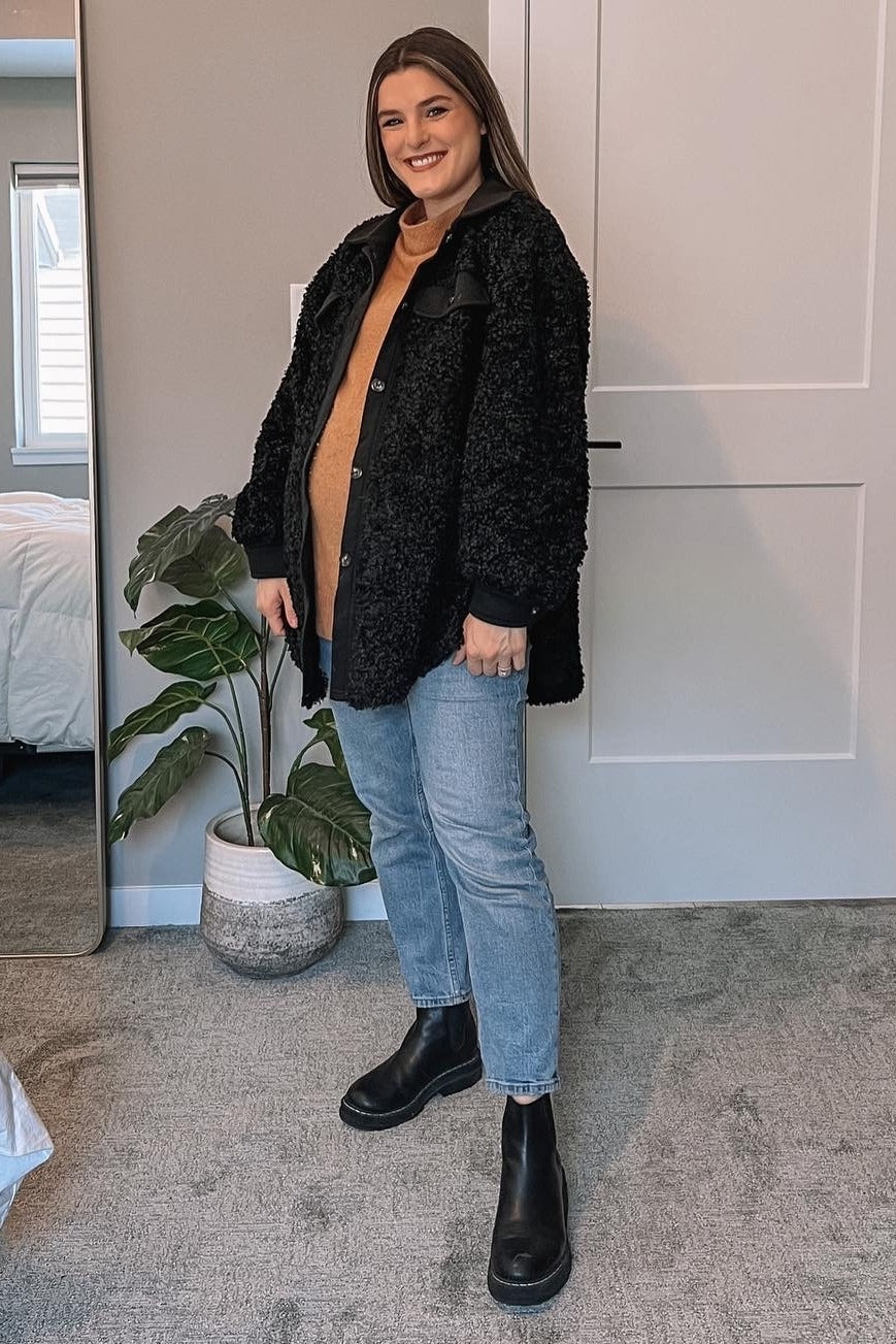 woman wearing black sherpa jacket, tan sweater, blue jeans, and black chelsea boots in a bedroom