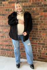 Smiling pregnant woman in black sherpa jacket, cream turtleneck, and distressed jeans standing against a brick wall