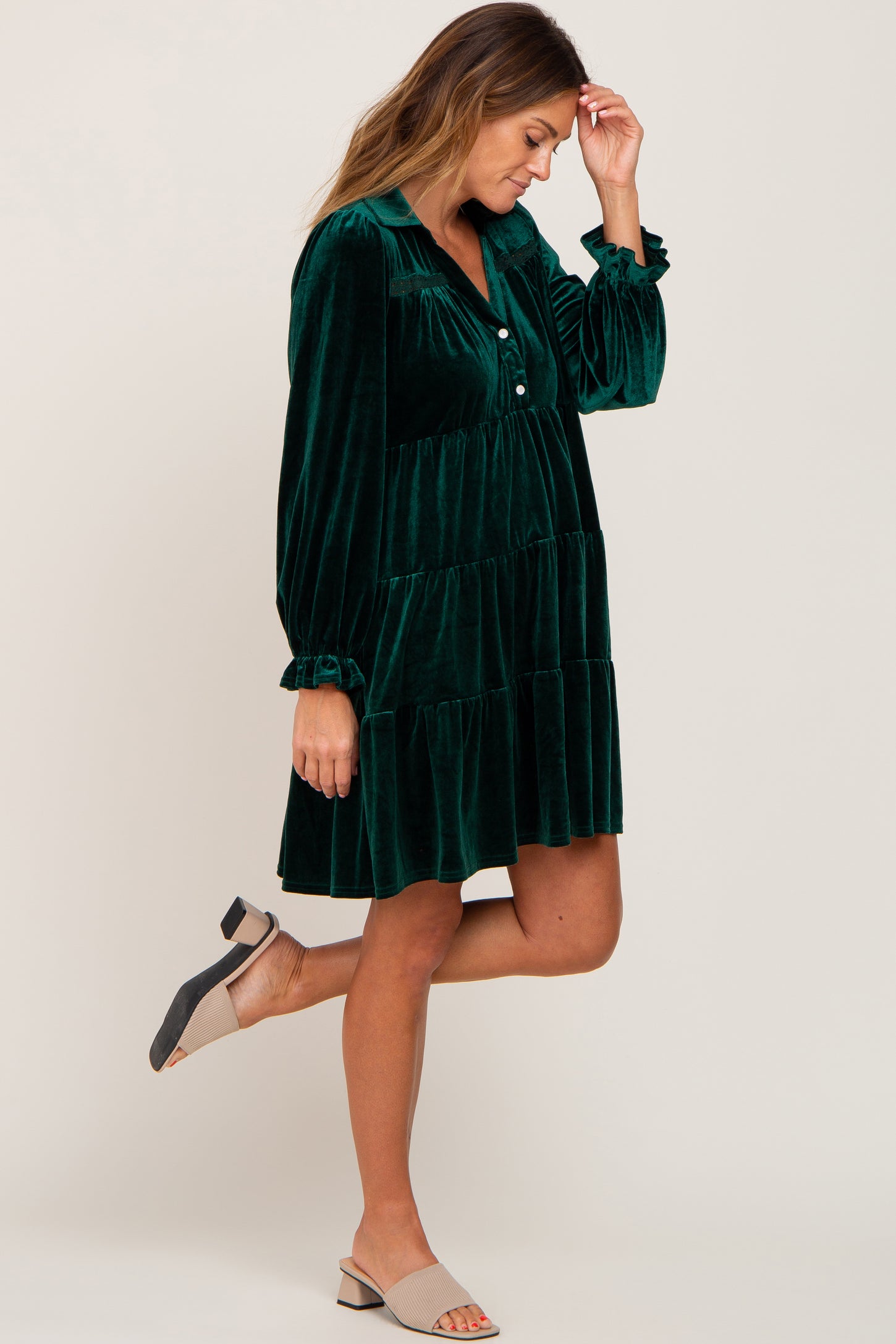 Woman wearing an emerald green velvet tiered maternity dress with long sleeves and tan block heel sandals