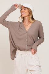 woman wearing a taupe textured button-up sweater with long sleeves and a relaxed fit, paired with light-colored pants and a cap