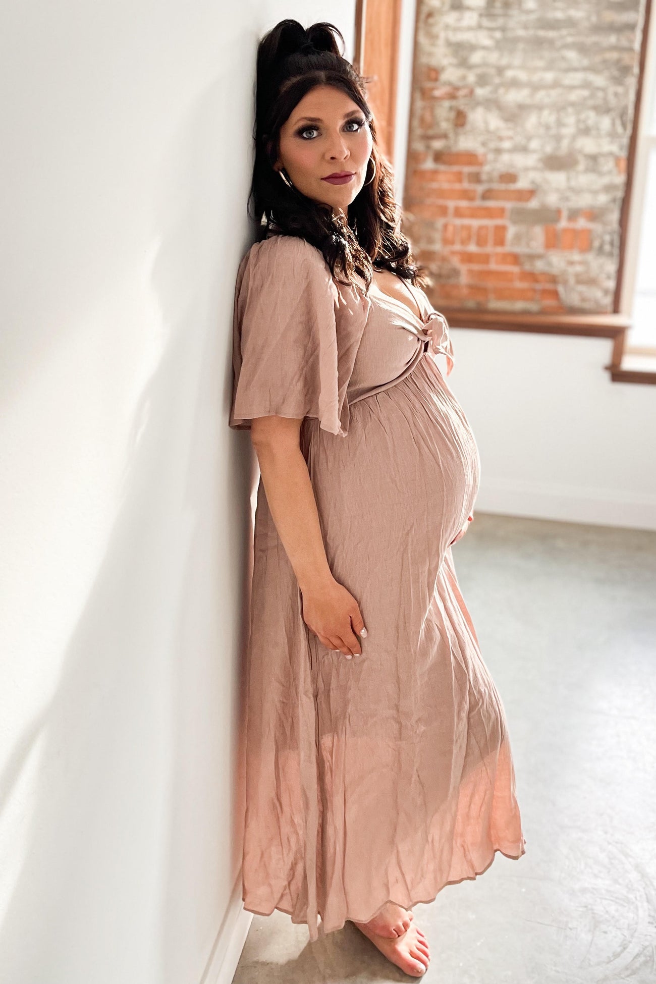woman wearing a blush pink maternity dress with short flutter sleeves, standing and showing her baby bump