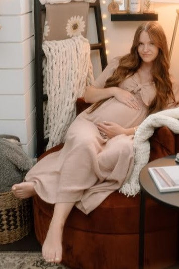 woman wearing a light beige maternity maxi dress with a bow detail, sitting in a cozy chair and showing her baby bump
