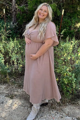 Smiling pregnant woman in a mauve maternity maxi dress and white cowboy boots posing outdoors