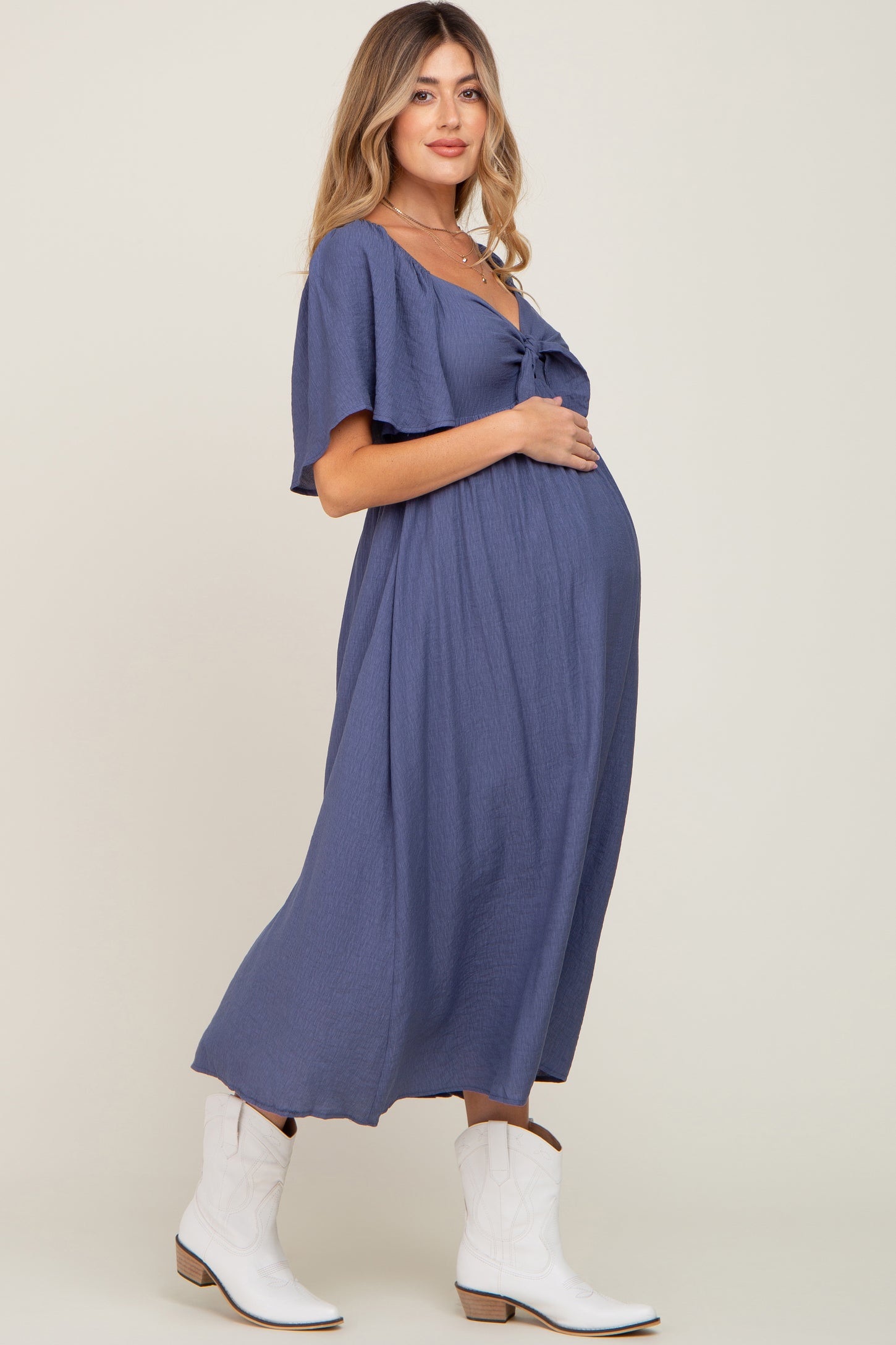 woman wearing a navy blue maternity dress with short sleeves and a tie front, showcasing her baby bump and wearing white boots