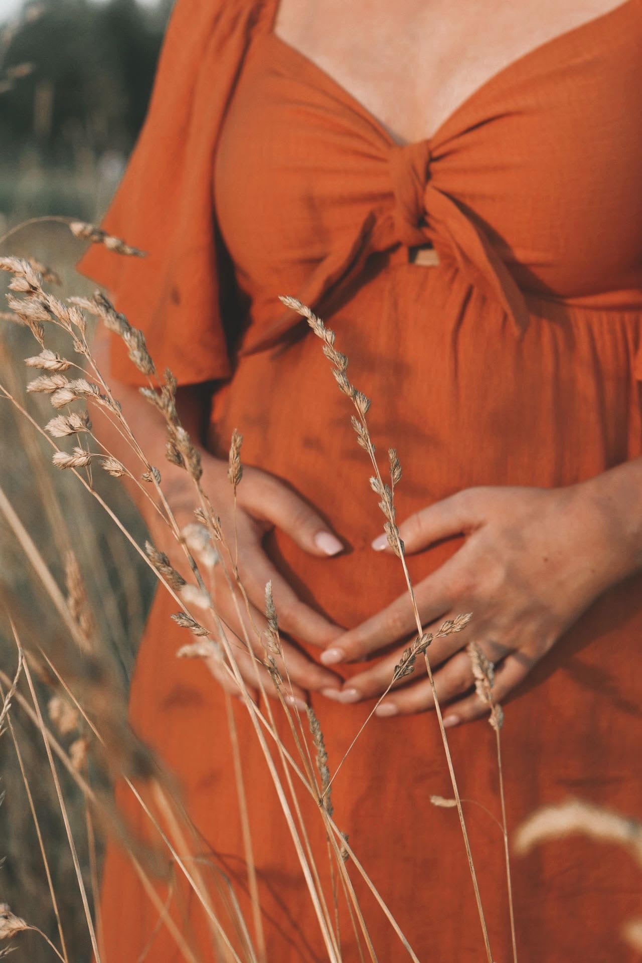 woman wearing an orange maternity dress with short sleeves and a tied front, holding her baby bump in a grassy field