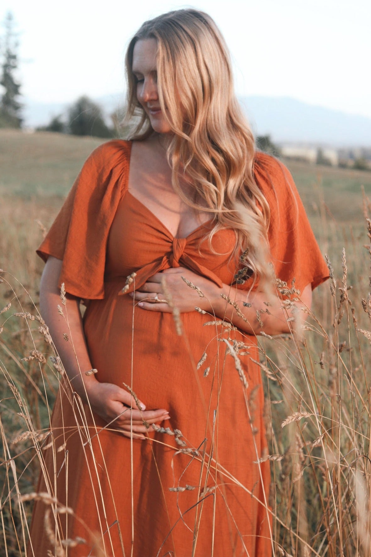 woman wearing an orange maternity dress with a knotted front, holding her baby bump in a field of tall grasses
