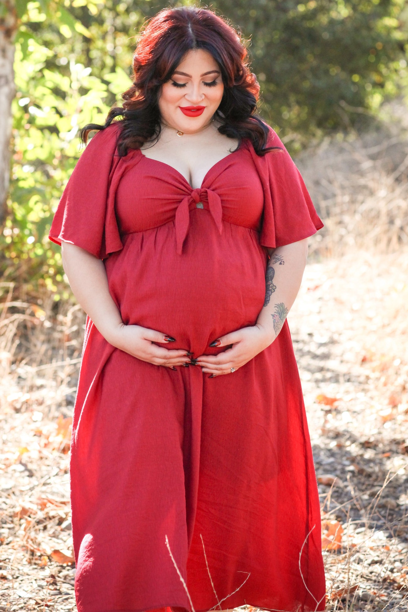 woman wearing a red maternity dress with a tie front detail, gently cradling her baby bump in an outdoor setting