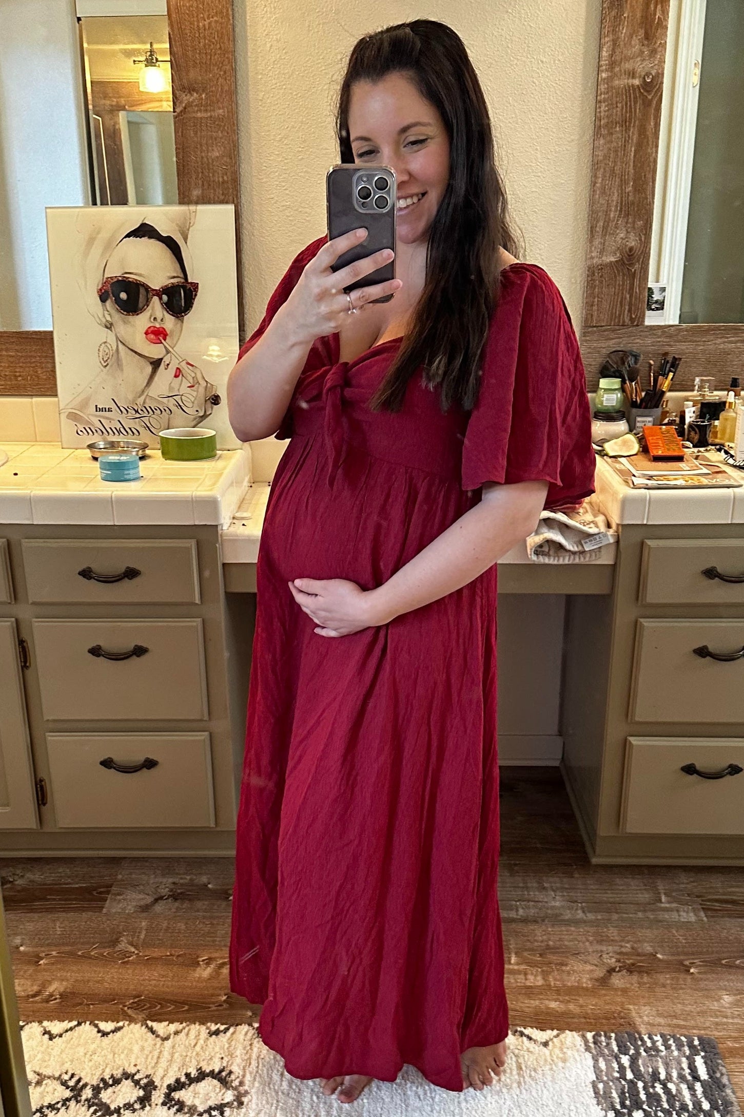 woman wearing a red maternity maxi dress with a bow detail, holding her baby bump in a bathroom mirror selfie